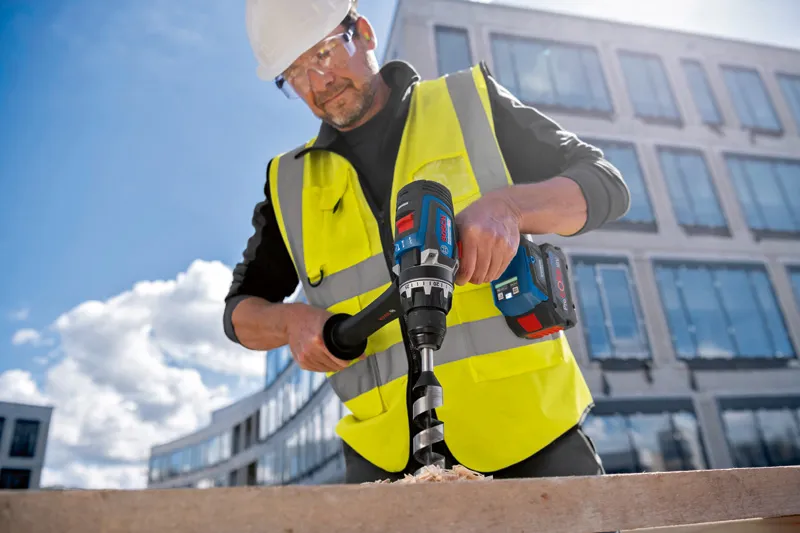 يقوم شخص يرتدي معدات السلامة بحفر ثقب كبير في الخشب باستخدام أداة لاسلكية.