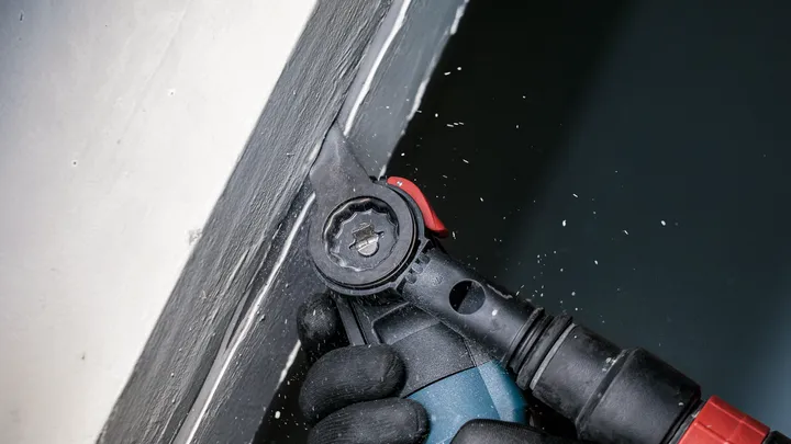 شخص يرتدي معدات السلامة يستخدم أداة كهربائية لصنفرة الطلاء بالقرب من زاوية الحائط.