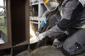 شخص يرتدي معدات السلامة يقوم بطحن شعاع معدني باستخدام مطحنة الزاوية.