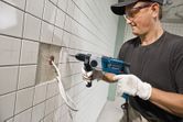 A person wearing safety equipment drills into a tiled wall near exposed wires.