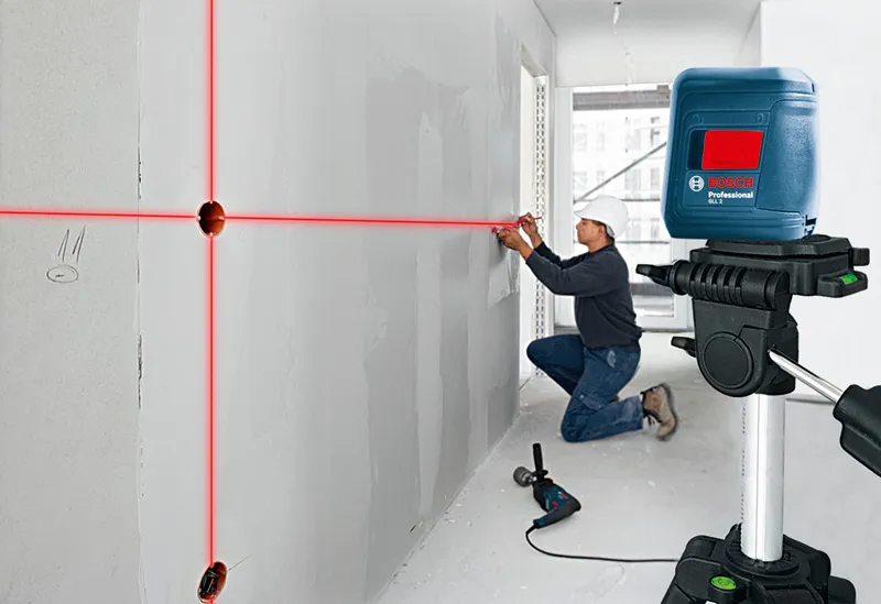 A person wearing safety equipment uses a laser leveling tool to mark lines on a wall.