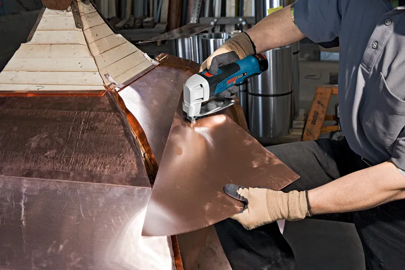 A person wearing safety equipment cuts sheet metal with a cordless metal shear.