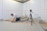 A person installs floor tiles in a bathroom, guided by a laser leveling tool on a tripod.