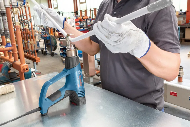 A person wearing safety equipment uses a heat gun to bend a plastic pipe.
