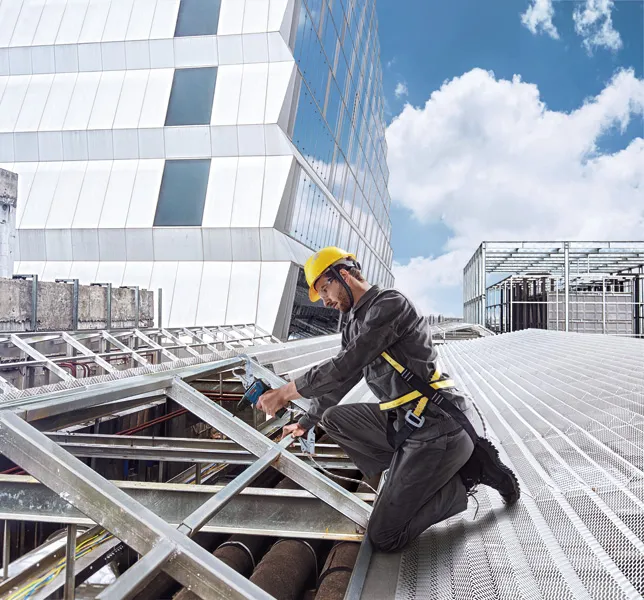 A person wearing safety equipment uses a cordless drill on a metal roof structure.
