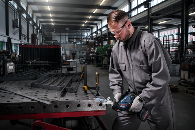 Person wearing safety equipment cuts metal on a workbench with a cordless saw.