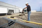 A person wearing safety equipment cuts a pipe with a cordless band saw on a rooftop.