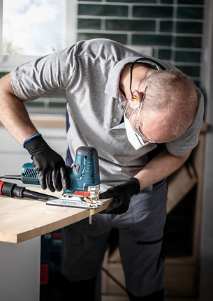 A person wearing safety equipment uses a jigsaw to cut a wooden board.
