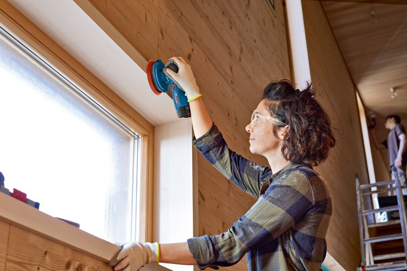 A person wearing safety equipment sands a wooden window frame with a cordless random orbit sander.