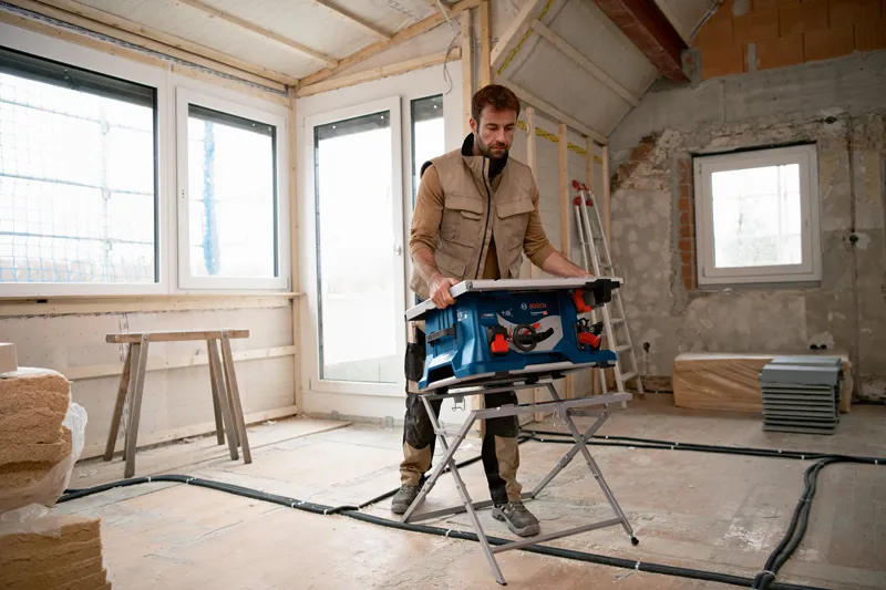 A person wearing safety equipment sets up a cordless table saw in a room under renovation.