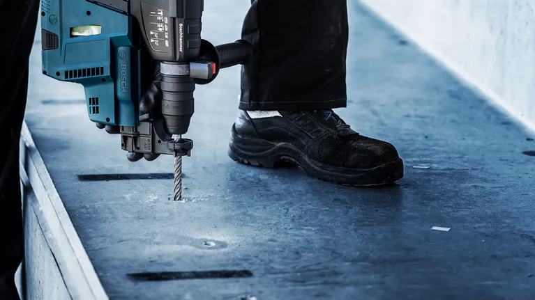 Person wearing safety equipment drills into concrete with a heavy-duty rotary hammer.