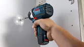 A person uses a cordless impact driver to drive a screw into a white surface.