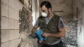 A person wearing safety equipment removes tiles from a wall using a power tool.