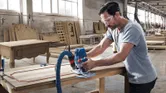A person wearing safety equipment shapes a wooden panel using a router in a workshop.