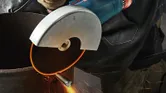A person wearing safety equipment cuts metal pipe with a handheld angle grinder.