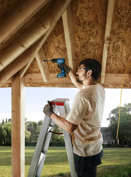 A person on a ladder drills into wooden rafters in a partially built structure.