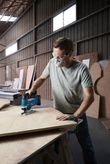 A person wearing safety equipment cuts plywood with a cordless jigsaw in a workshop.