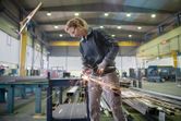 A person wearing safety equipment uses an angle grinder to cut metal in a factory.