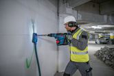 A person wearing safety equipment drills into a concrete wall with a cordless rotary hammer.