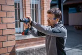 A person wearing safety equipment drills into a brick wall with a cordless power drill.
