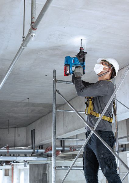 A person wearing safety equipment drills into a ceiling while standing on scaffolding.