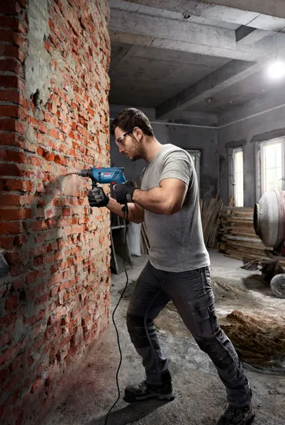 A person wearing safety equipment drills into a brick wall with an impact drill.