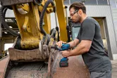 A person wearing safety equipment uses an impact wrench on heavy machinery outdoors.