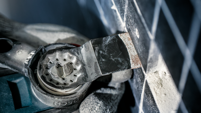 Person wearing safety equipment removes grout from tile joints with a multi-tool.