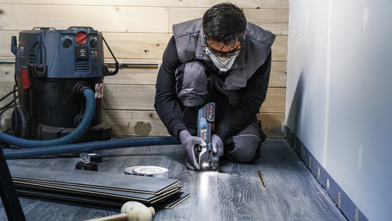 A person wearing safety equipment cuts laminate flooring with a power tool.