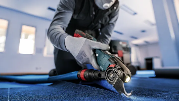 Person wearing safety equipment cuts flooring with a power tool in a brightly lit room.