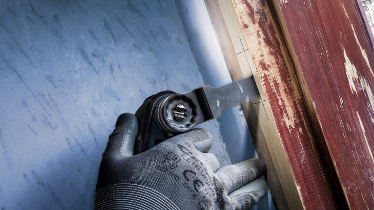 Person wearing safety equipment uses a multi-tool to cut wood near a painted surface.
