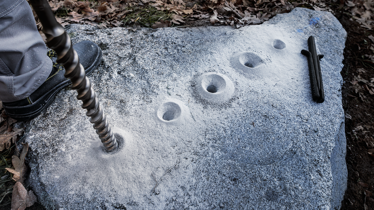 Person drilling multiple holes into a large stone outdoors.