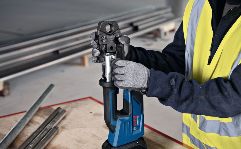 A person wearing safety equipment uses a cordless press tool at a workbench.