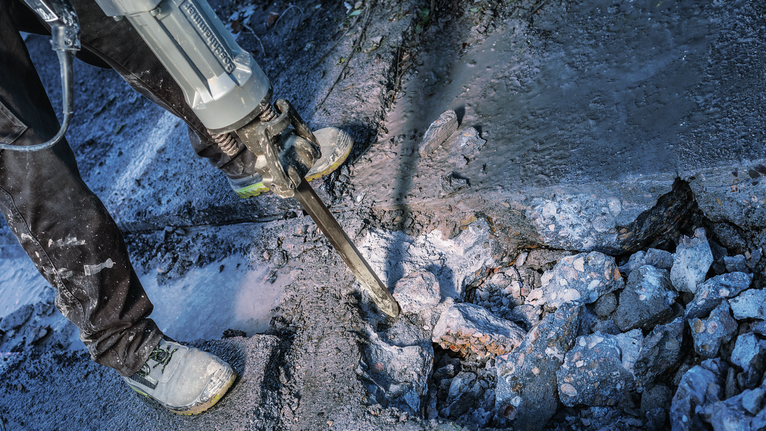 Person wearing safety equipment breaks concrete with a demolition hammer.