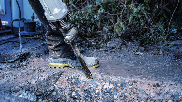 Person wearing safety equipment uses a demolition hammer to break wet concrete.
