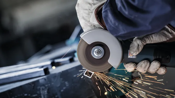Person wearing safety equipment uses an angle grinder to cut metal, creating sparks.
