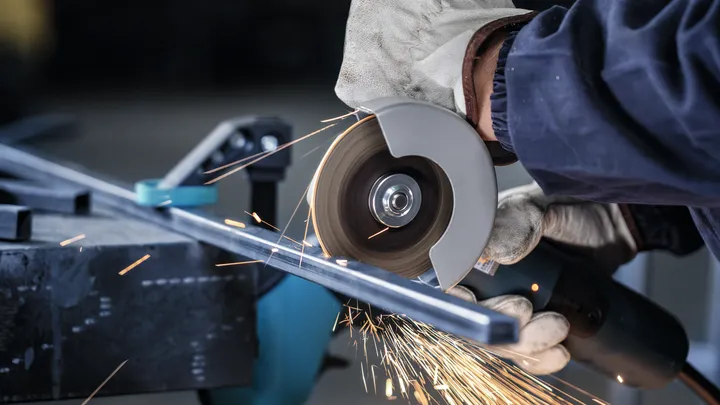 Person wearing safety equipment cuts metal with a grinder, creating sparks.