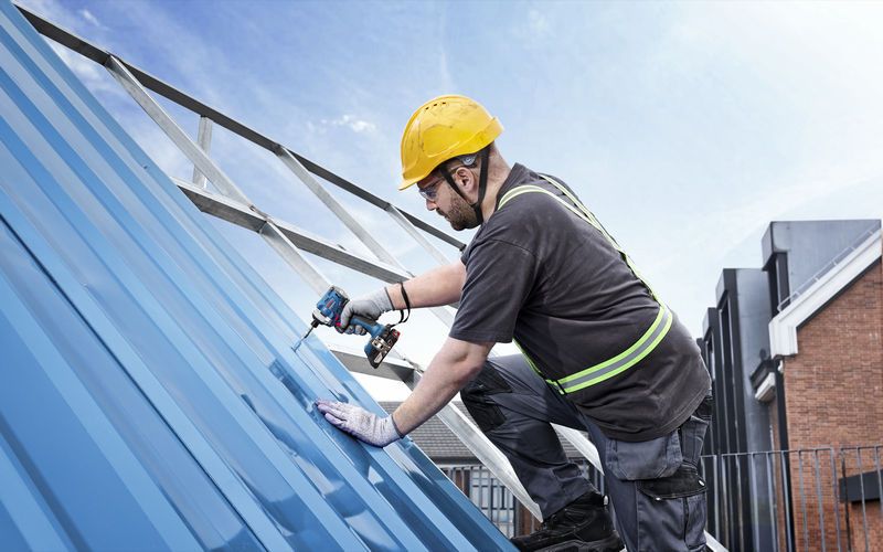 A person wearing safety equipment drives a screw into a metal roof with a cordless drill.