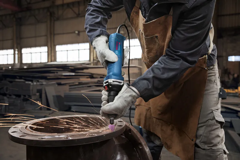A person wearing safety equipment grinds metal with a straight grinder, producing sparks.