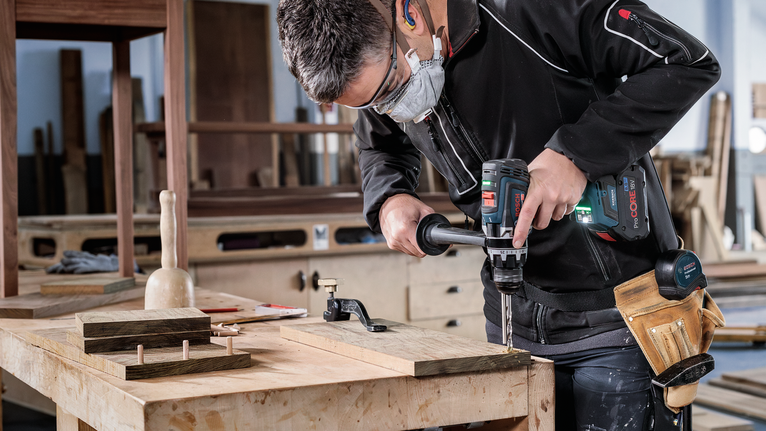 Person wearing safety equipment drills into a wooden board on a workbench.