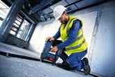 A person wearing safety equipment uses a power tool to drill into a concrete floor.