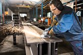 A person wearing safety equipment grinds a steel beam in an industrial workshop.