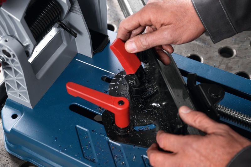 A person adjusts a metal cut-off saw angle, shavings cover the blue base.