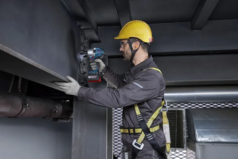 A person wearing safety equipment tightens bolts on a steel beam using a cordless impact wrench.