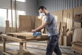 A person wearing safety equipment planes a thick wooden slab in a carpentry workshop.
