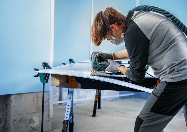 A worker wearing safety equipment cuts a white panel using a jigsaw.