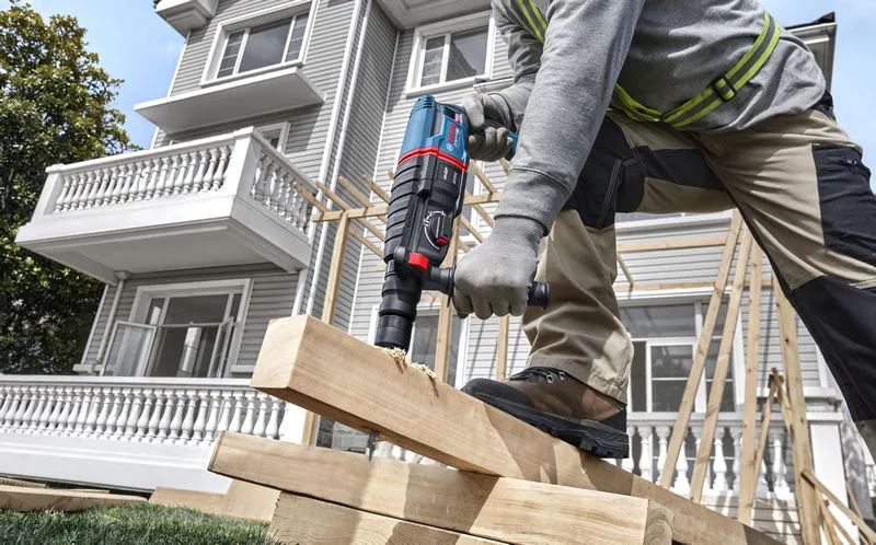 A person wearing safety equipment drills into stacked wooden beams at a construction site.