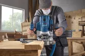 A person wearing safety equipment uses a router to shape the edge of a wooden board.