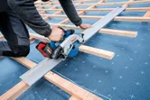 A person using a circular saw to cut a metal sheet on a rooftop frame.