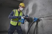 A person wearing safety equipment uses a demolition hammer to break a concrete wall.